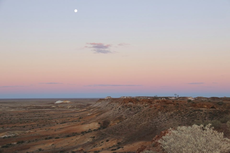 A long ridge of eroded red-brown hills above a wide desert plain under a pink and lilac dusk sky, a full moon high above