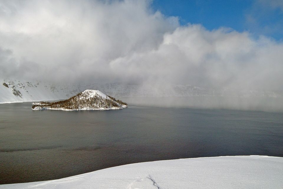 A small forested island dusted with snow on dark grey water under a great bank of cloud, with a smooth slope of snow in the foreground