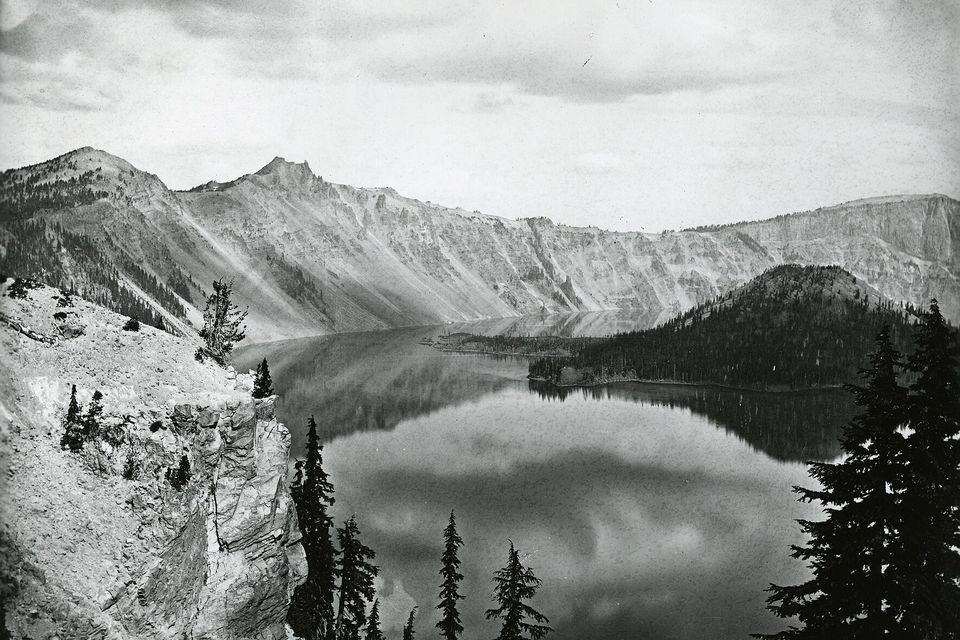 A black-and-white print of a still lake reflecting cloud, steep pale rim walls behind, a conifer-covered island at right and a rocky bluff at left