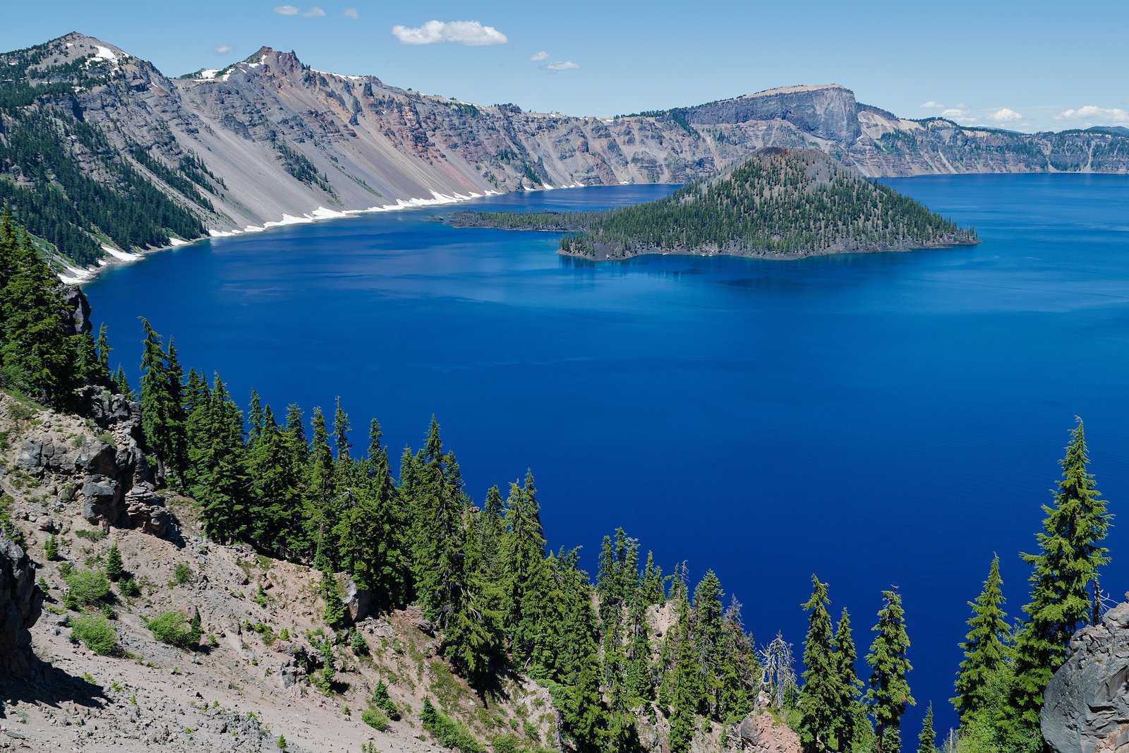 A deep blue crater lake ringed by steep pale walls, a forested cinder cone rising from the water, patches of snow along the far rim and dark conifers on the slope in the foreground