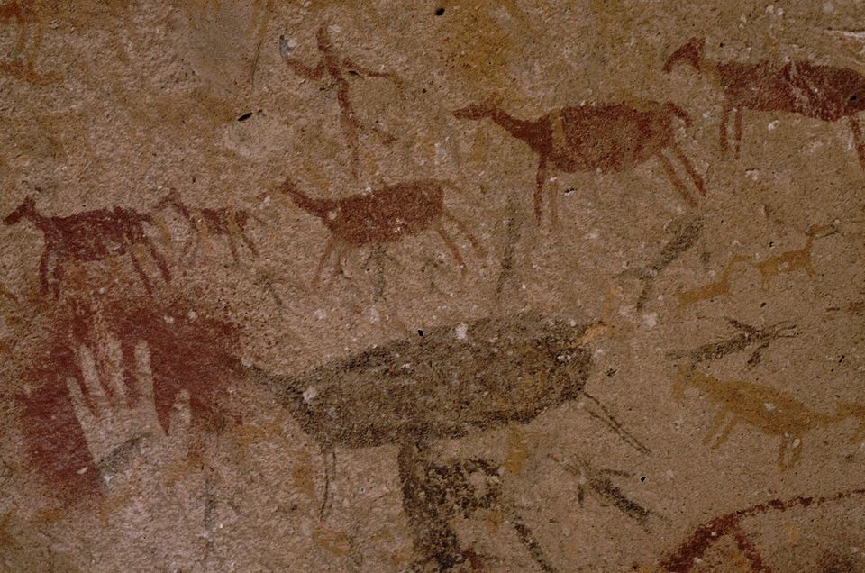 Painted guanacos and small figures in red and ochre on pale rock, with a faint hand stencil at lower left