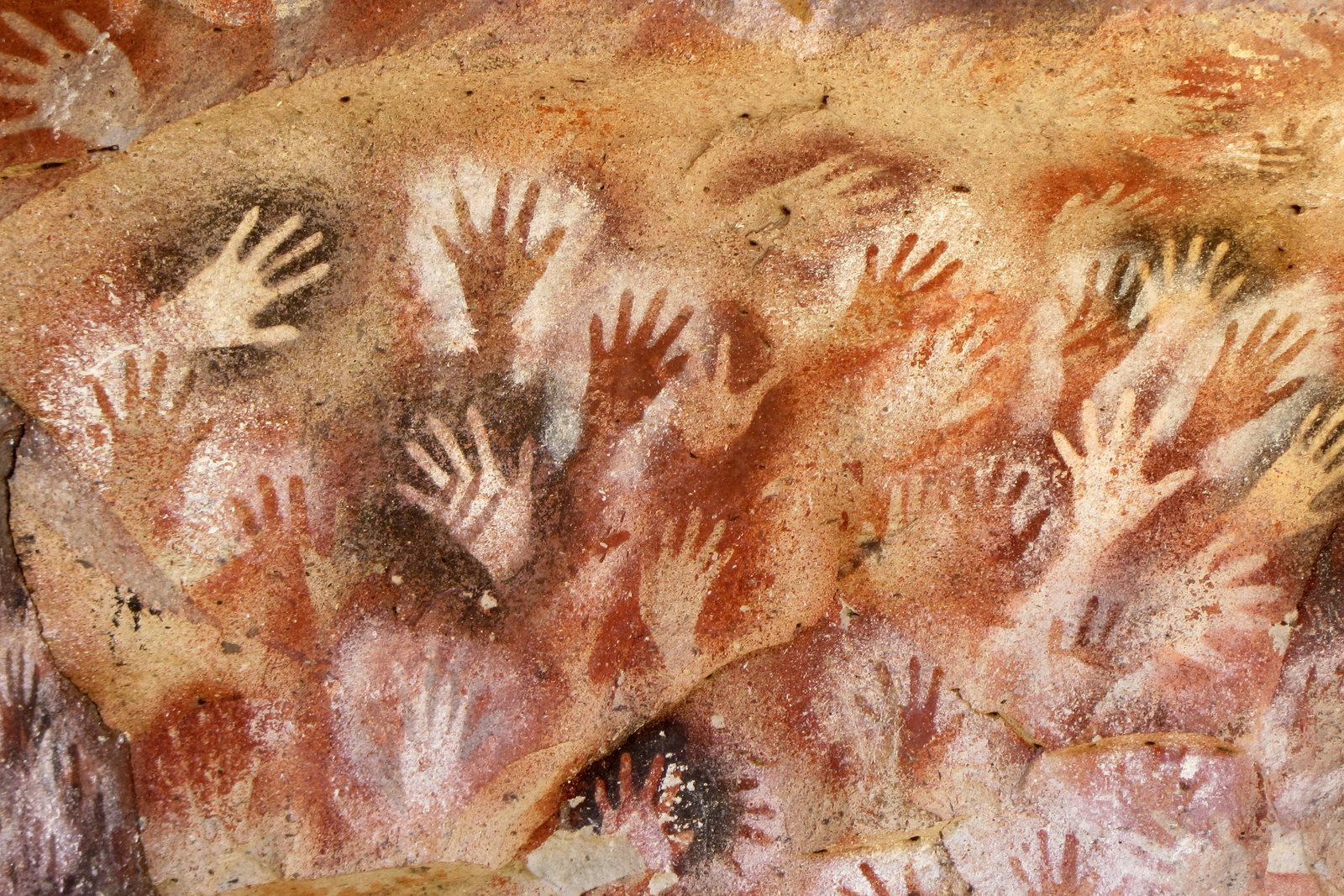 A rock face covered with dozens of stencilled human hands in red, ochre and white, overlapping one another across the stone