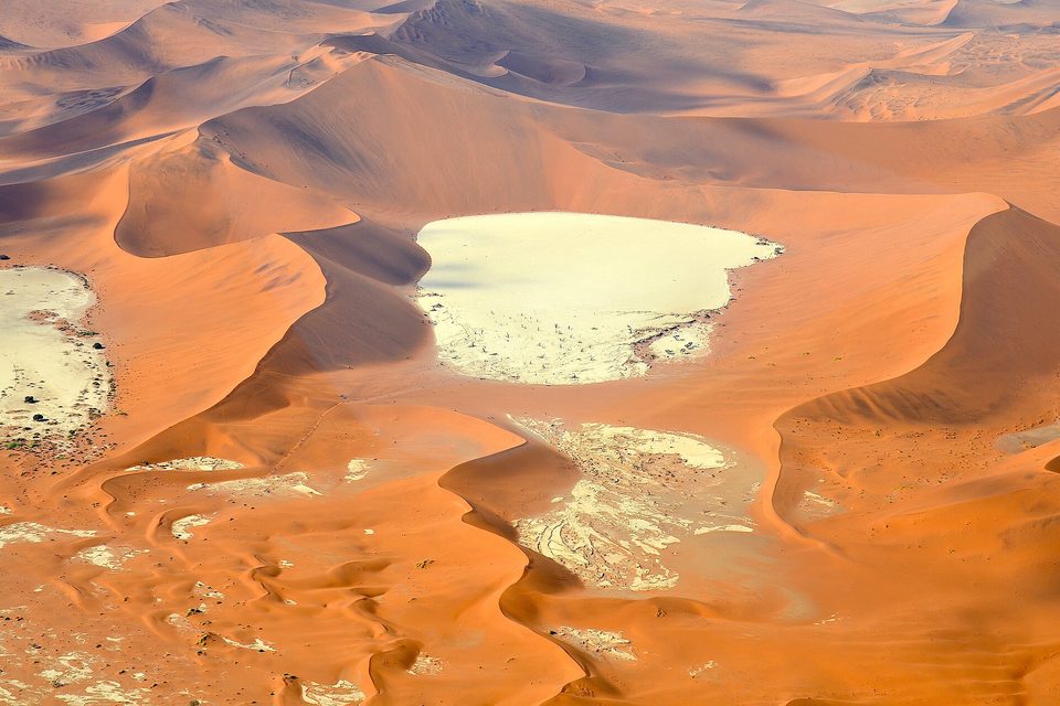An aerial view of orange sand dunes in curving ridges, with a flat white clay pan enclosed among them and a second smaller pan below