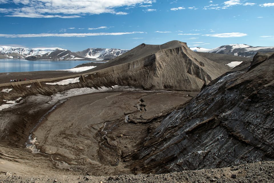 A landscape of bare brown volcanic ash and eroded ridges with patches of old snow, the flooded caldera bay and white mountains in the distance