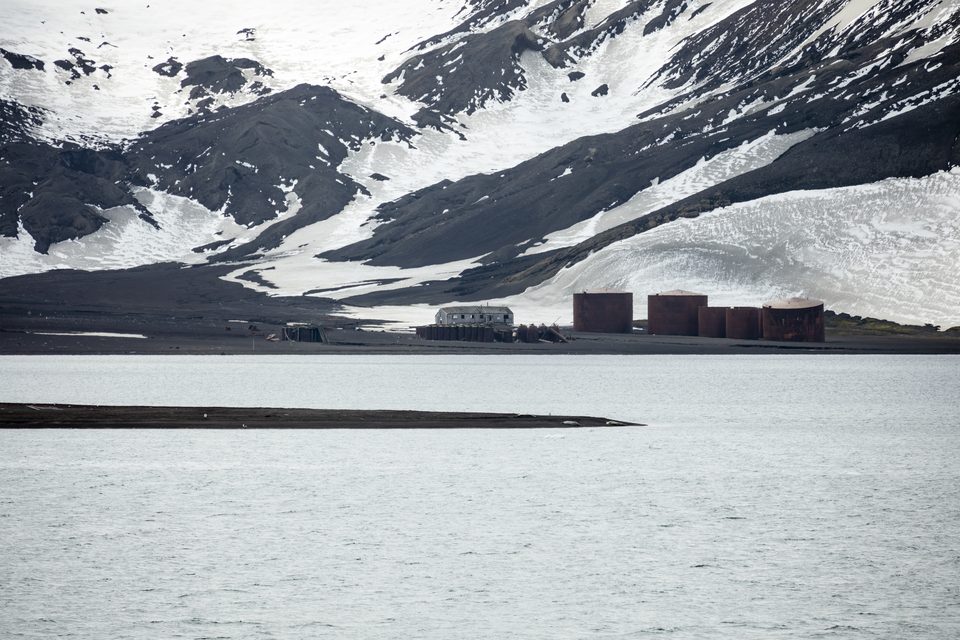 Rust-red whaling-station tanks and a weathered shed on a black-sand shore, snow-streaked ash hills behind