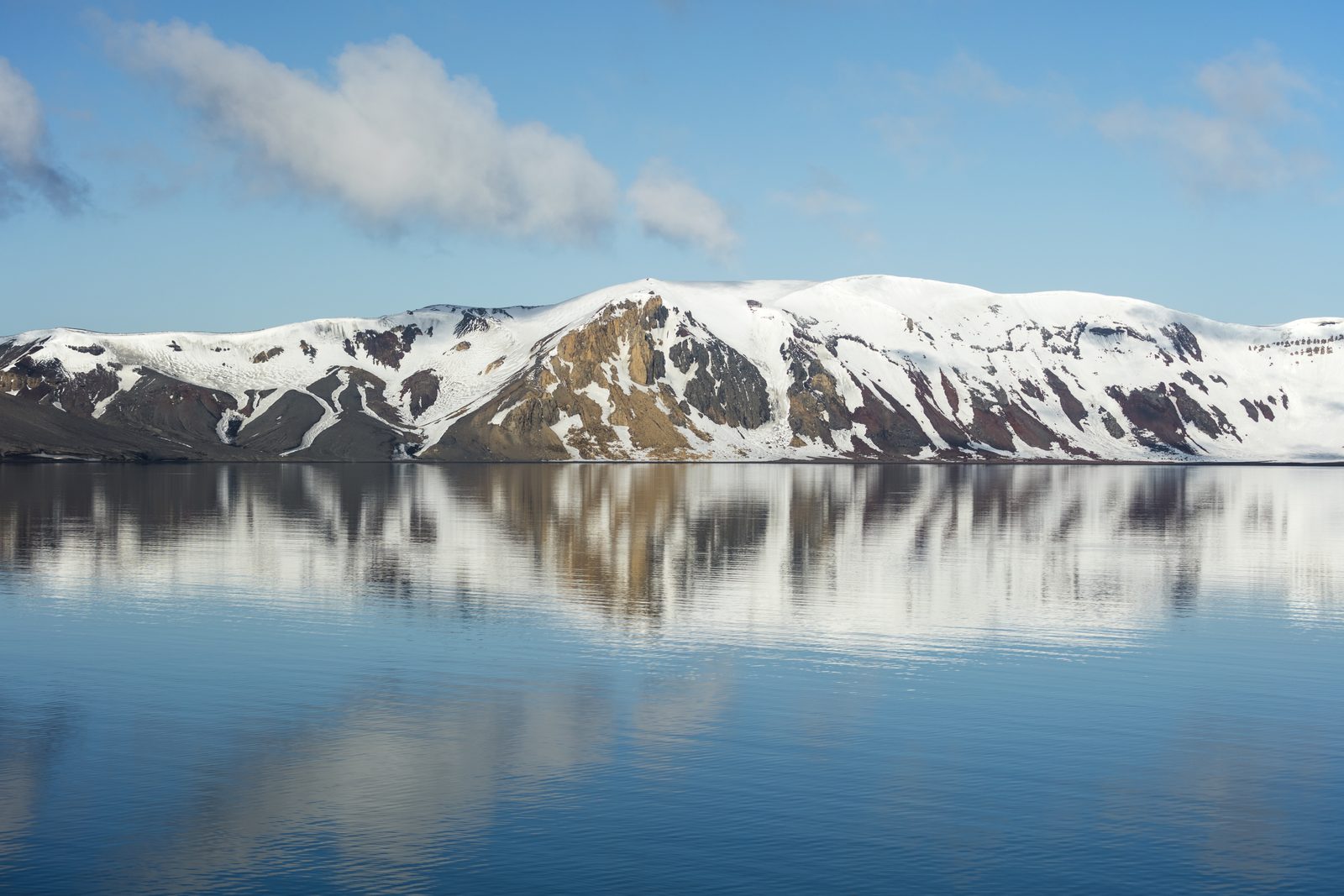 A snow-streaked red and ochre volcanic crater wall mirrored in perfectly still water under a blue sky with a few clouds
