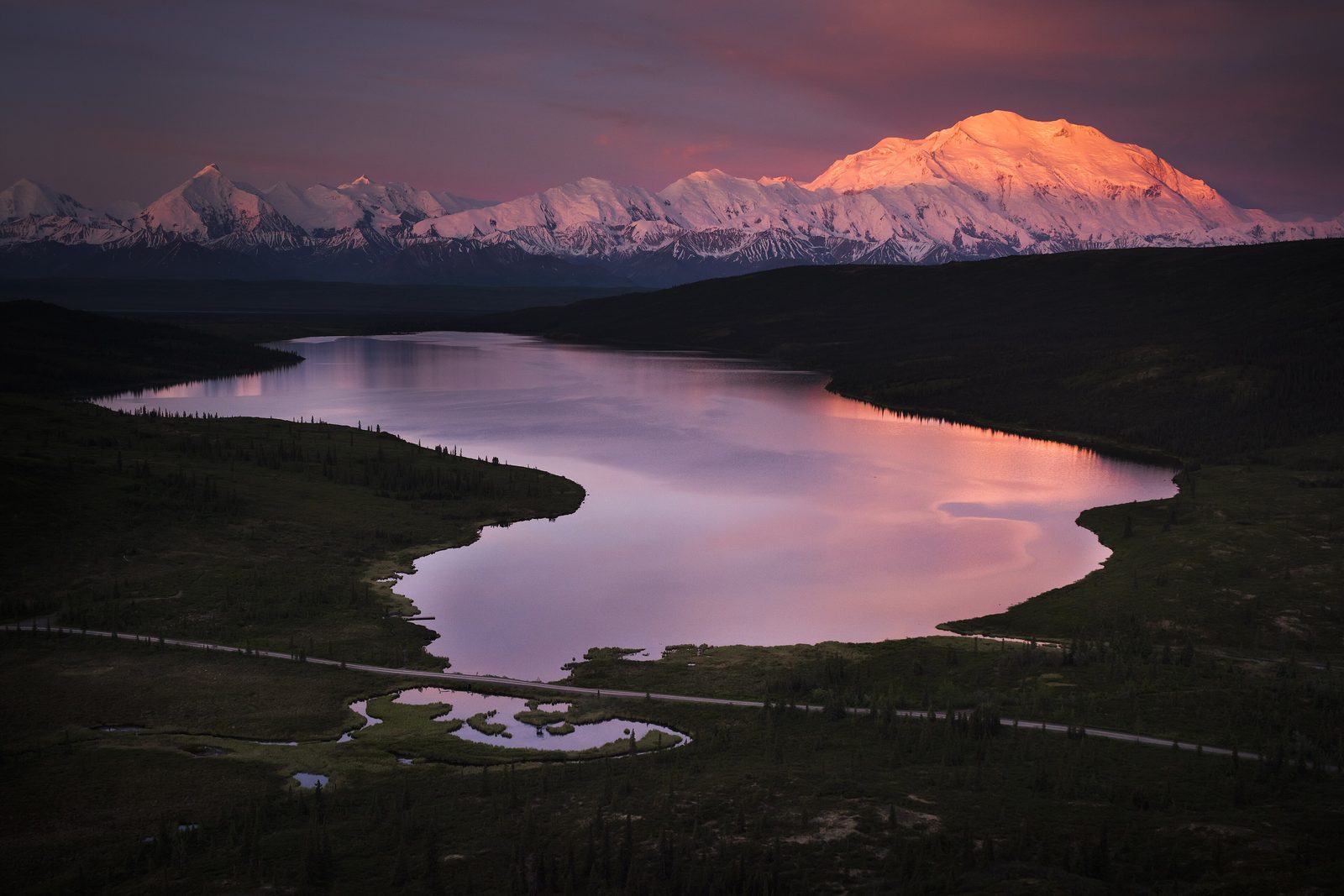 A huge snow-covered mountain lit pink at dawn, above dark tundra hills and a still lake reflecting the sky