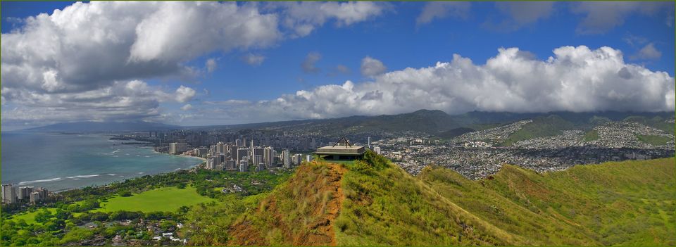 A wide view from a crater rim over a concrete roof, a green park, a city of towers and the coast
