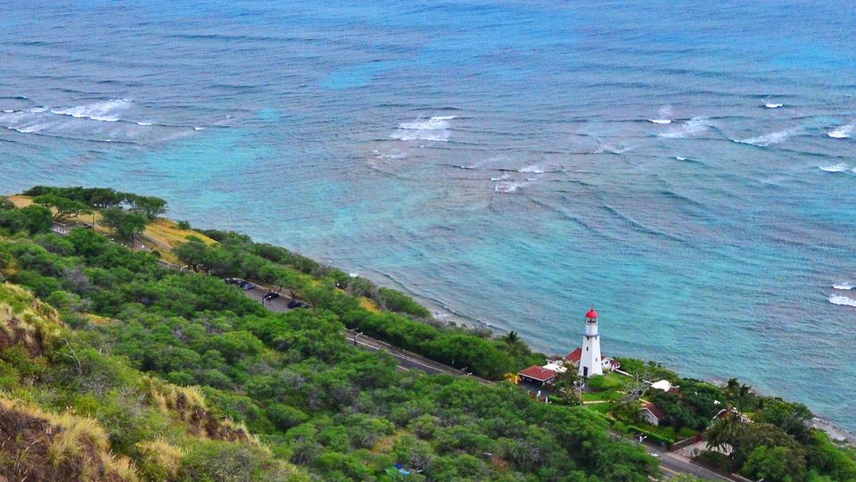 A white lighthouse tower with red roofs on a green slope above a turquoise reef