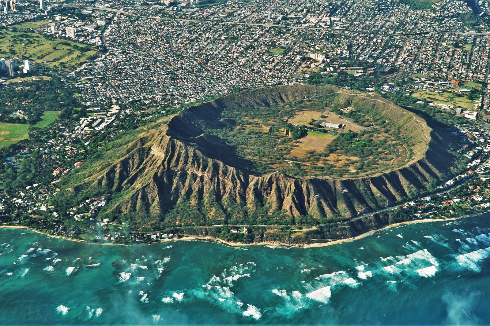 An aerial view of a brown volcanic cone with a grassy crater floor, green park land below and a turquoise reef with white surf in the foreground