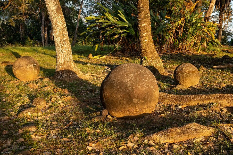 A smooth round stone sphere glows orange in low sun on a root-crossed, pebbly ground, two more spheres and palm trunks behind