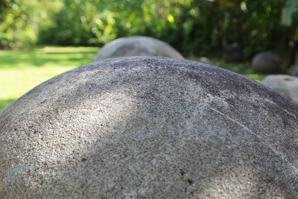 A close view across the curved, speckled grey surface of a large stone sphere, more spheres soft-focused on a green lawn behind