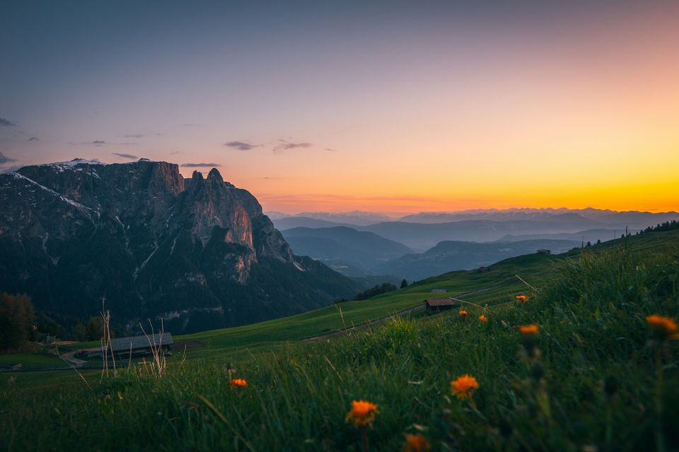 A high flowering meadow with a hay hut beneath a dark rock massif and an orange sky