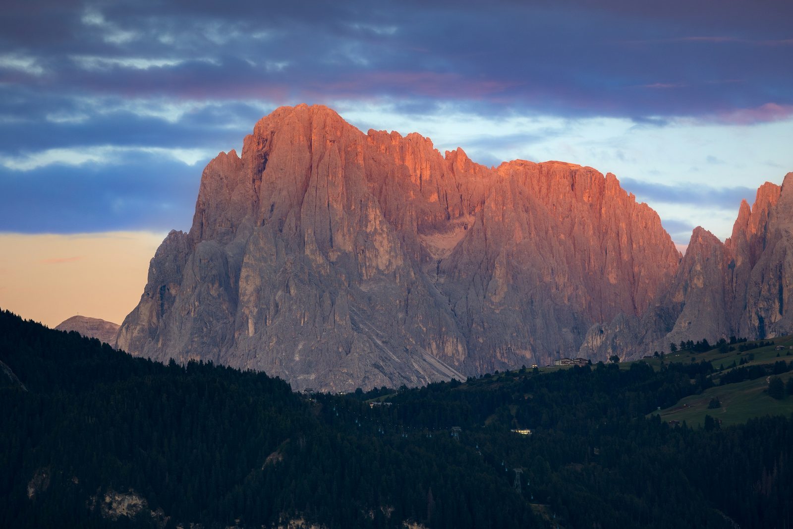 A great pale rock wall lit pink above dark forest under a blue evening sky
