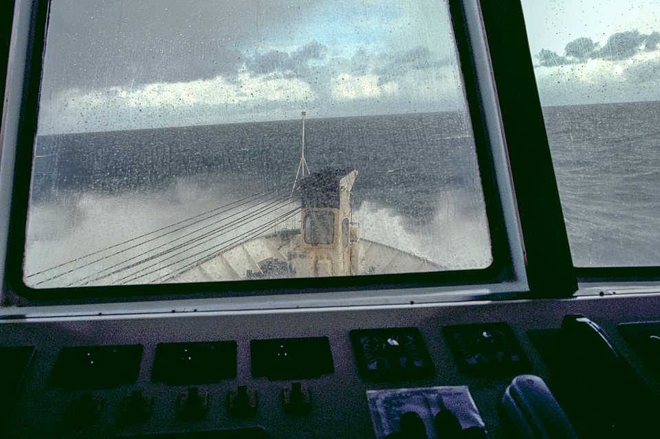 A ship's bow throwing white spray into a grey sea, seen through rain-spotted bridge windows above a control console