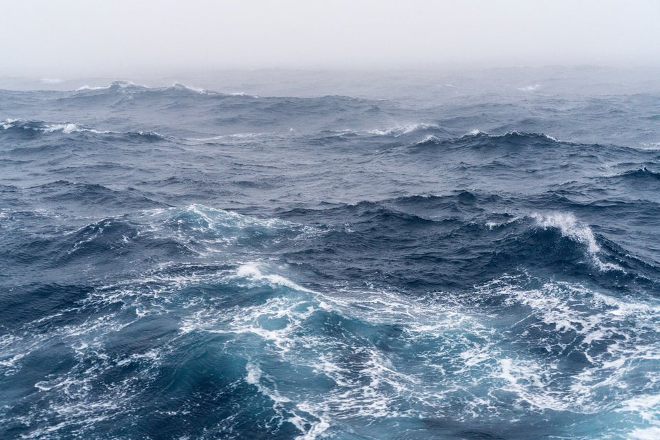 Rows of steep dark-blue waves with breaking white crests running to a horizon lost in grey fog
