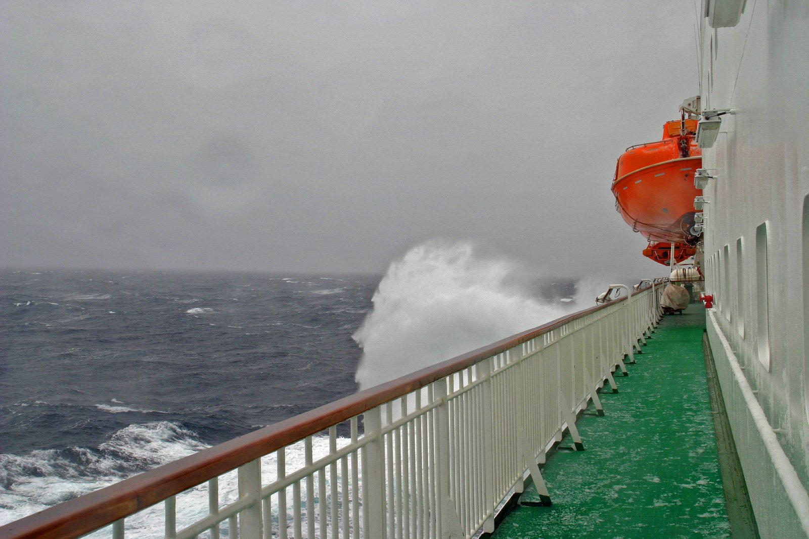 A wave breaking against the side of a ship, spray thrown above the rail, grey open sea and low cloud beyond, orange lifeboat on the deck