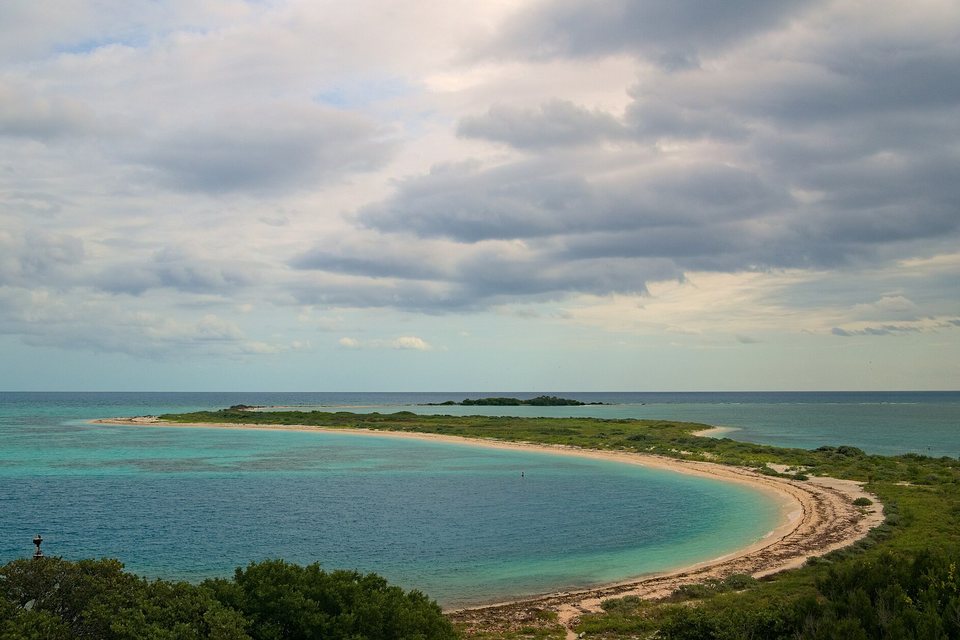 A long curving sand spit fringed with low green scrub around a pale turquoise lagoon, under a broken grey sky