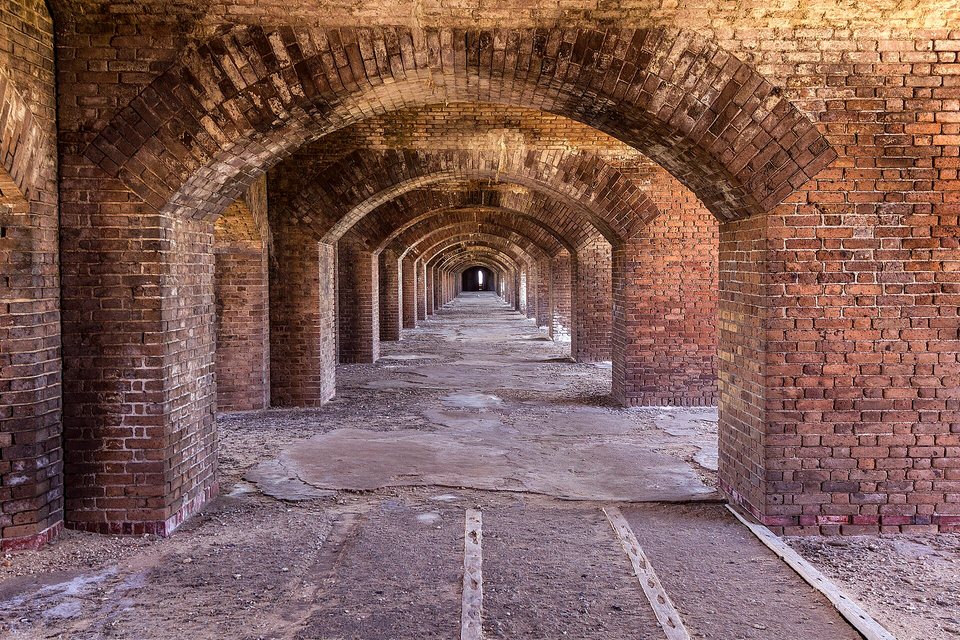 A long corridor of repeating red-brick arches receding to a small bright opening, a worn stone floor below
