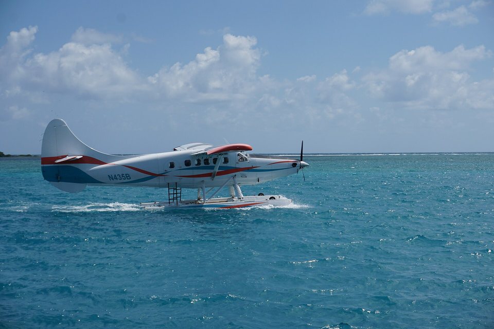 A white floatplane with red and blue stripes skimming across turquoise water, spray at its floats, a low reef line behind