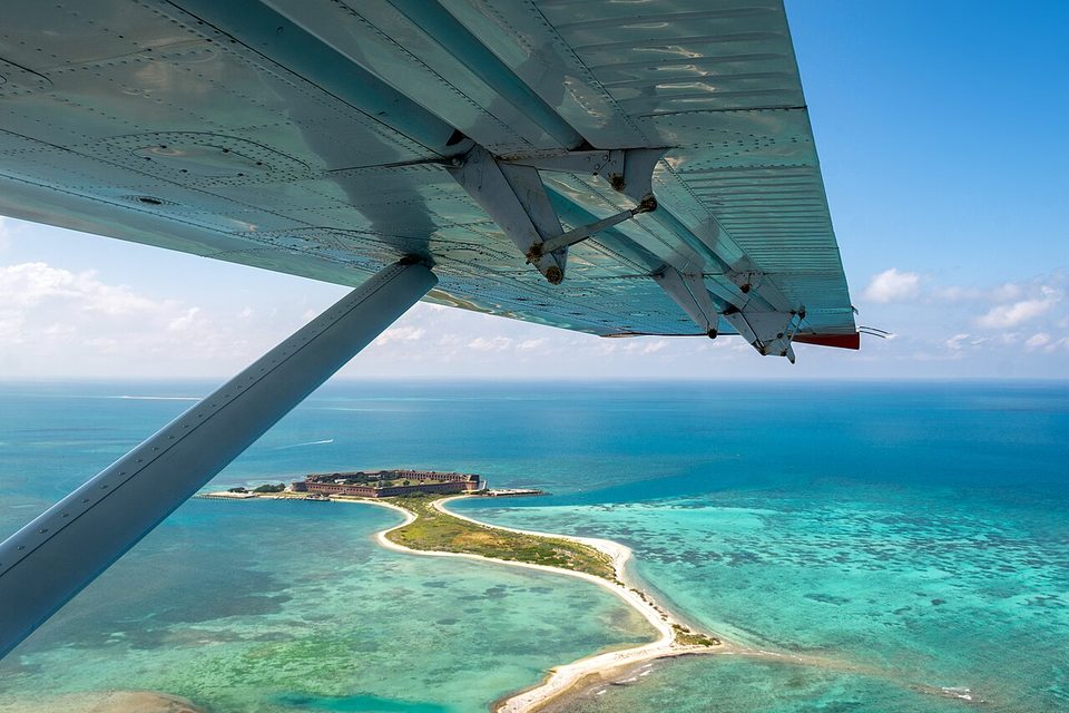 Fort Jefferson and the Bush Key sand spit seen from the air beneath a plane's wing, a turquoise reef and shallow water all around