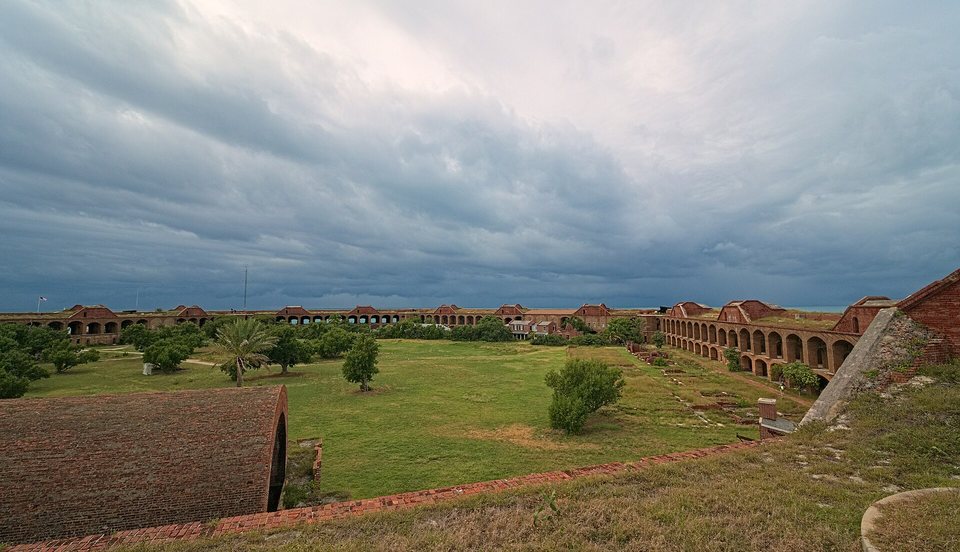 A wide grassy parade ground with palms and trees enclosed by brick arcades, under heavy dark storm clouds