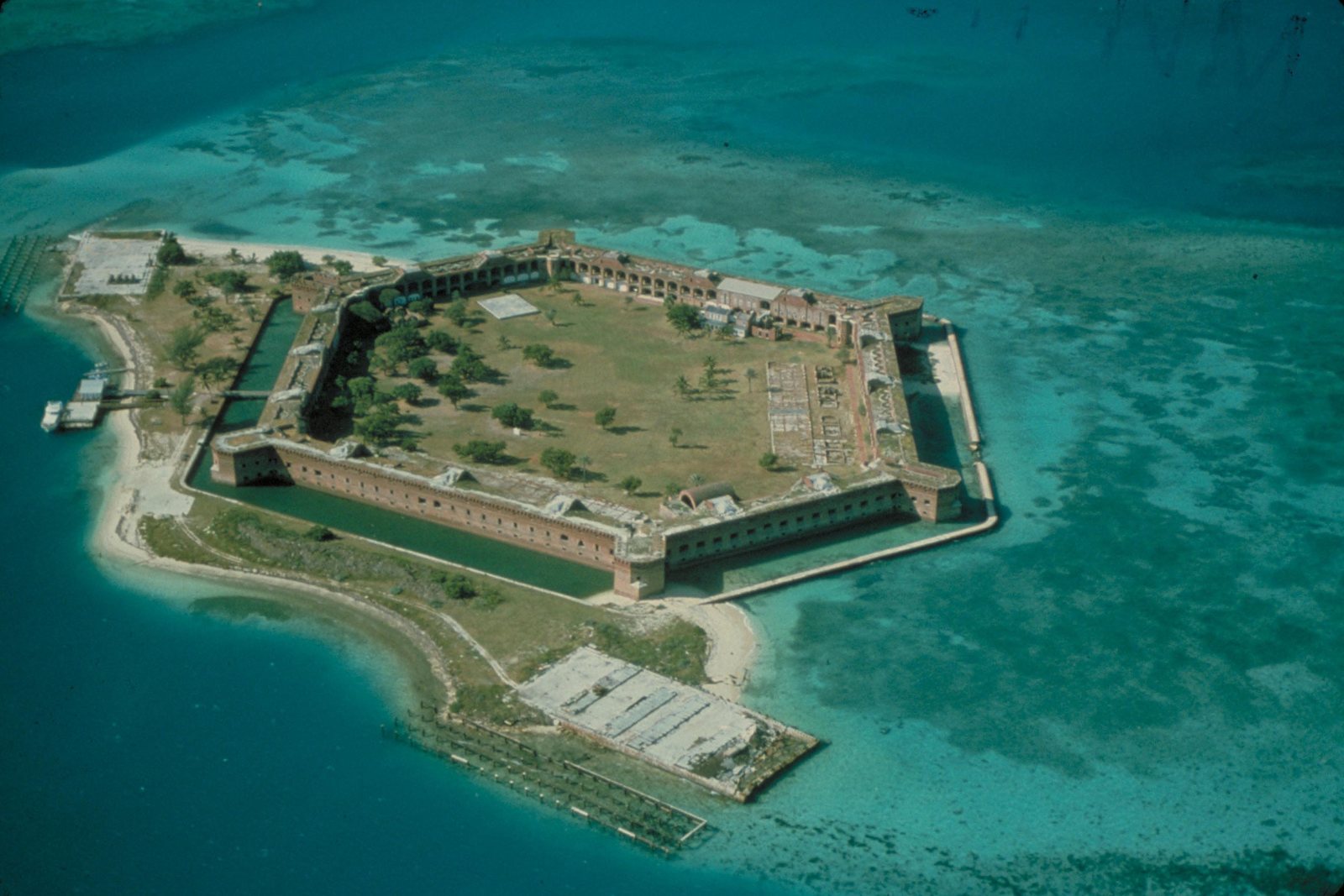 Aerial view of a six-sided brick fortress with a moat, occupying almost all of a small key surrounded by turquoise shallows and reef