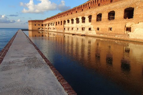 A long brick fort wall with rows of arched openings lit warm orange, reflected in the still moat beside a narrow walkway, blue sky and white cloud above