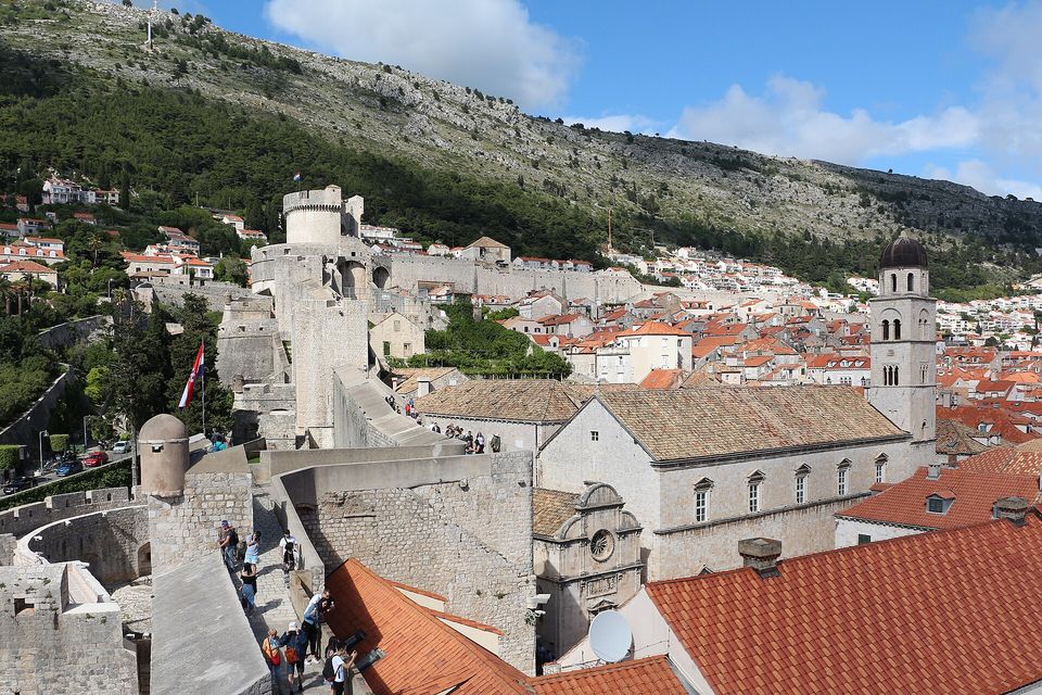 A stone wall walk running toward a round tower and a bell tower, a wooded ridge rising behind