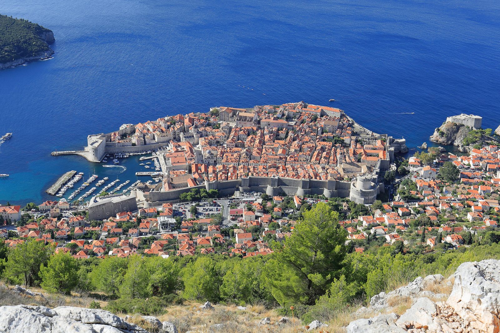 A walled harbour city of red roofs on a rocky point, an island offshore and a fortress on a rock at the right, in clear morning light