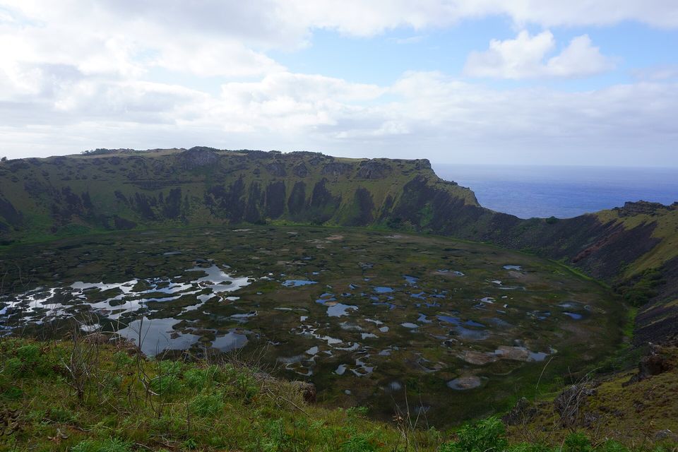 A wide volcanic crater with a floor of reeds and scattered pools of open water, green walls dropping to it and a strip of sea beyond the far rim under cloud
