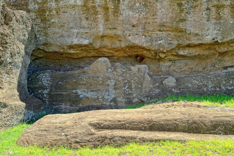 A long unfinished figure carved in relief and still attached to a pale lichen-spotted rock wall, a worked stone block lying on the grass in front