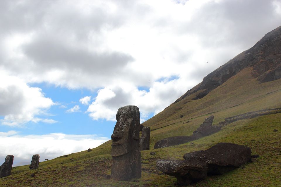 A tall moai in profile on a green slope with two more standing at the left and fallen figures lying on the grass at the right, cloud and blue sky above a dark rock ridge