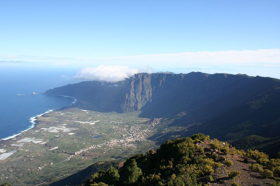 A vast curved cliff wall drops to a green coastal plain dotted with farms and villages, with deep-blue ocean at left and cloud on the rim