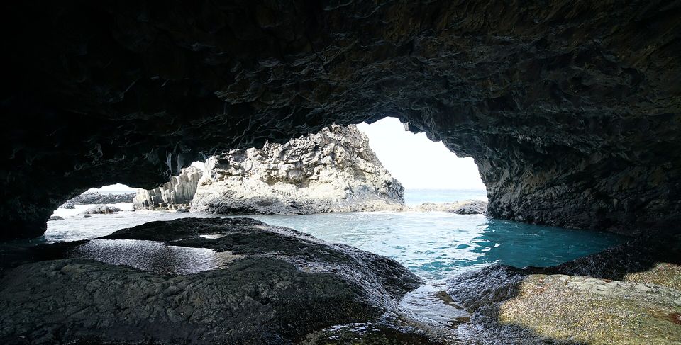 From inside a dark basalt cave, a natural arch frames a turquoise pool, pale rocks and the open sea