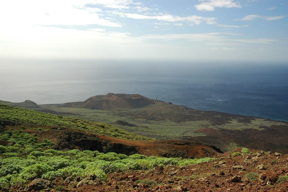 A rust-red volcanic cone stands on a dark lava headland above a calm sea, a small white lighthouse beside it, with green spurge scrub in the foreground