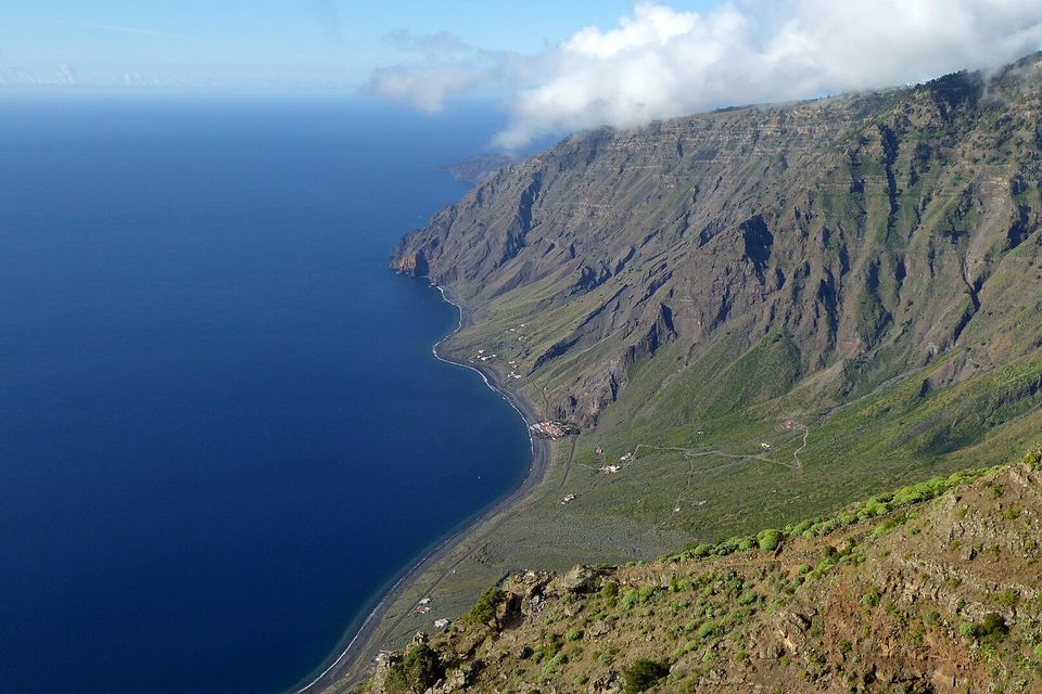A long sweep of ribbed cliff curves down to a dark beach and a deep-blue bay, cloud resting on the clifftop