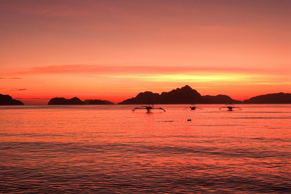 Three outrigger boats and a line of dark islands in silhouette under a red and orange sunset sky over Bacuit Bay