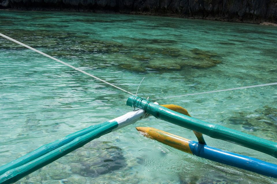 The bamboo outrigger of a boat resting over clear turquoise water and coral in Bacuit Bay