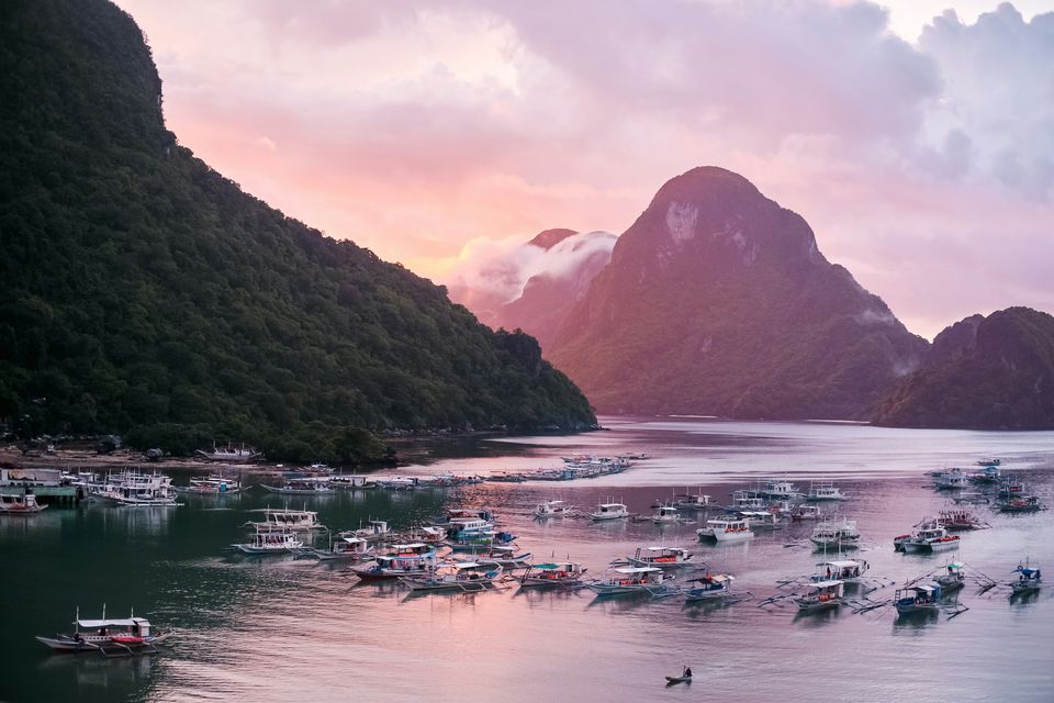 Outrigger boats moored in El Nido's bay at dusk, under a steep forested cliff and a pink sky