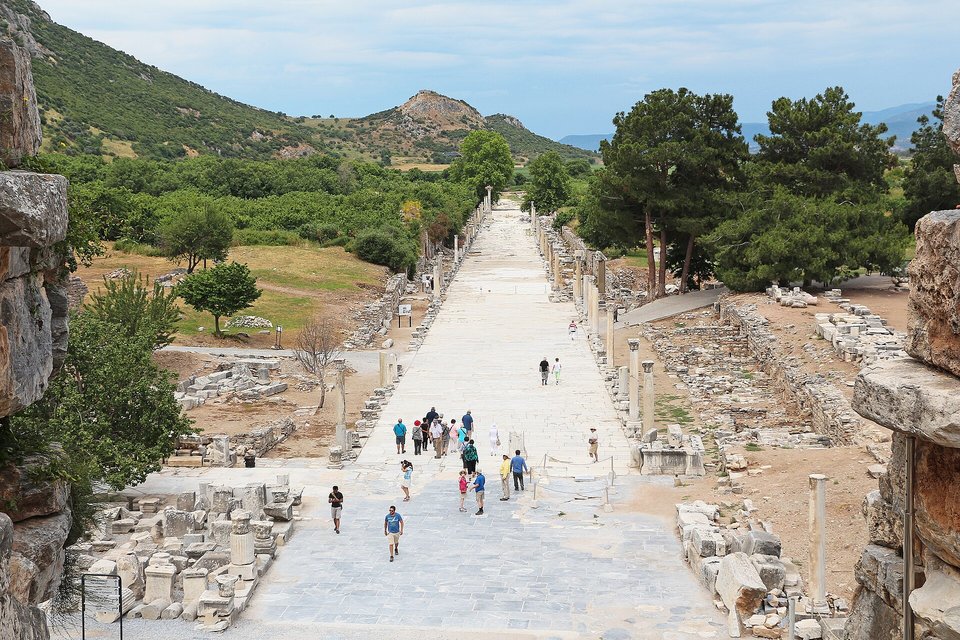 A long pale marble street lined with broken columns running away across a green plain toward low hills, small groups of visitors walking along it