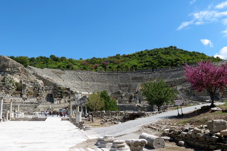 A great curved theatre of stone seats on a wooded hillside beyond a pale paved area and a gravel path, a flowering pink tree at the right