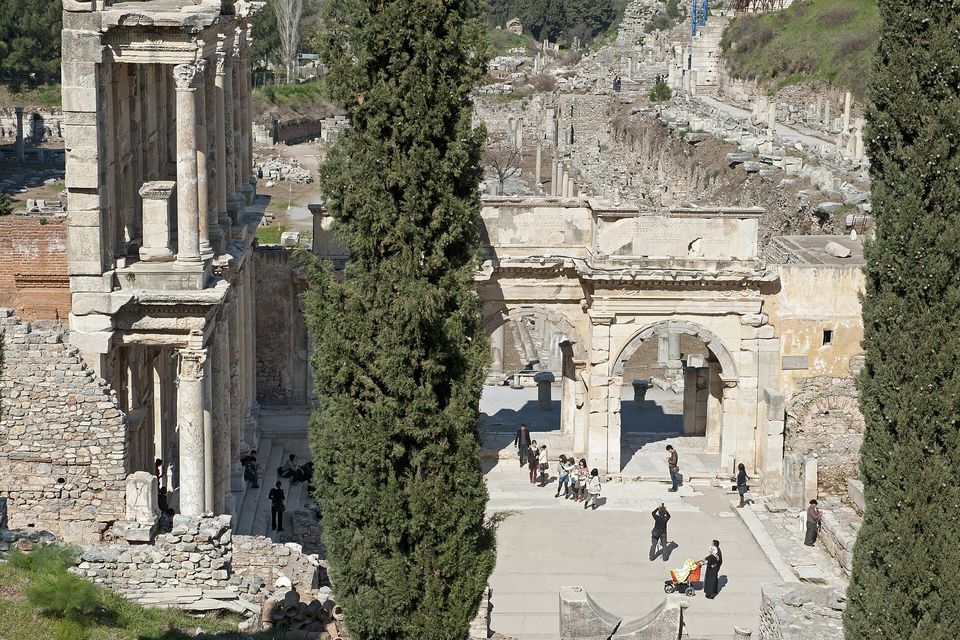 The corner of a Roman library façade beside a monumental gate, seen from above between two dark cypress trees, with tiny visitors on the paving