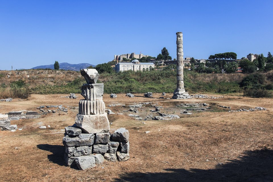 A tall column of stacked stone drums standing in a dry sunken field scattered with fallen blocks, a domed mosque and a hilltop castle in the distance under blue sky