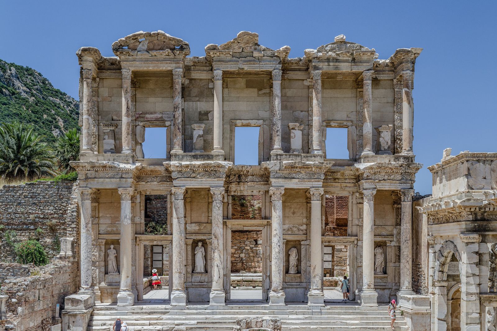 The two-storey stone façade of a Roman library with pairs of columns, empty window openings and niches, framed by palms and a blue sky