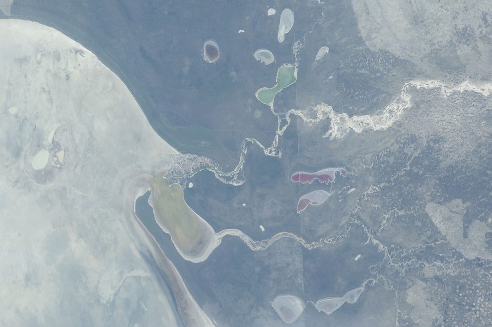 View from orbit of a pale salt pan edge, a winding river and small coloured pools in grey-brown country