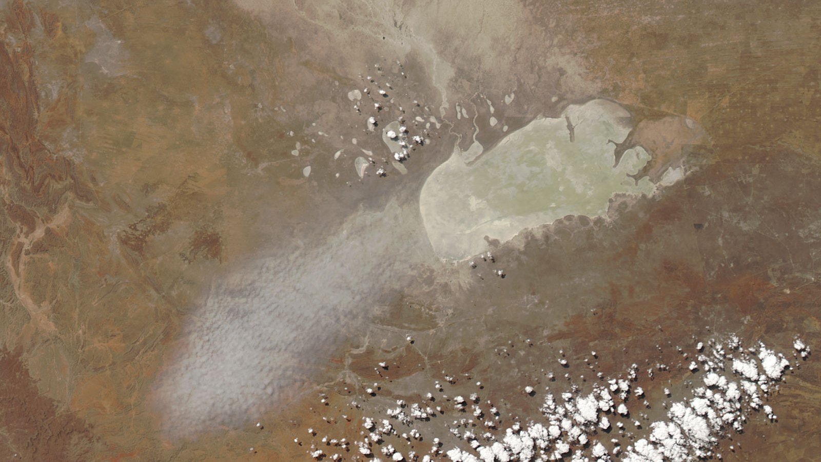 A satellite view of a pale oval salt pan in dry brown savannah, a plume of white dust streaming off its western shore across the land to the south-west