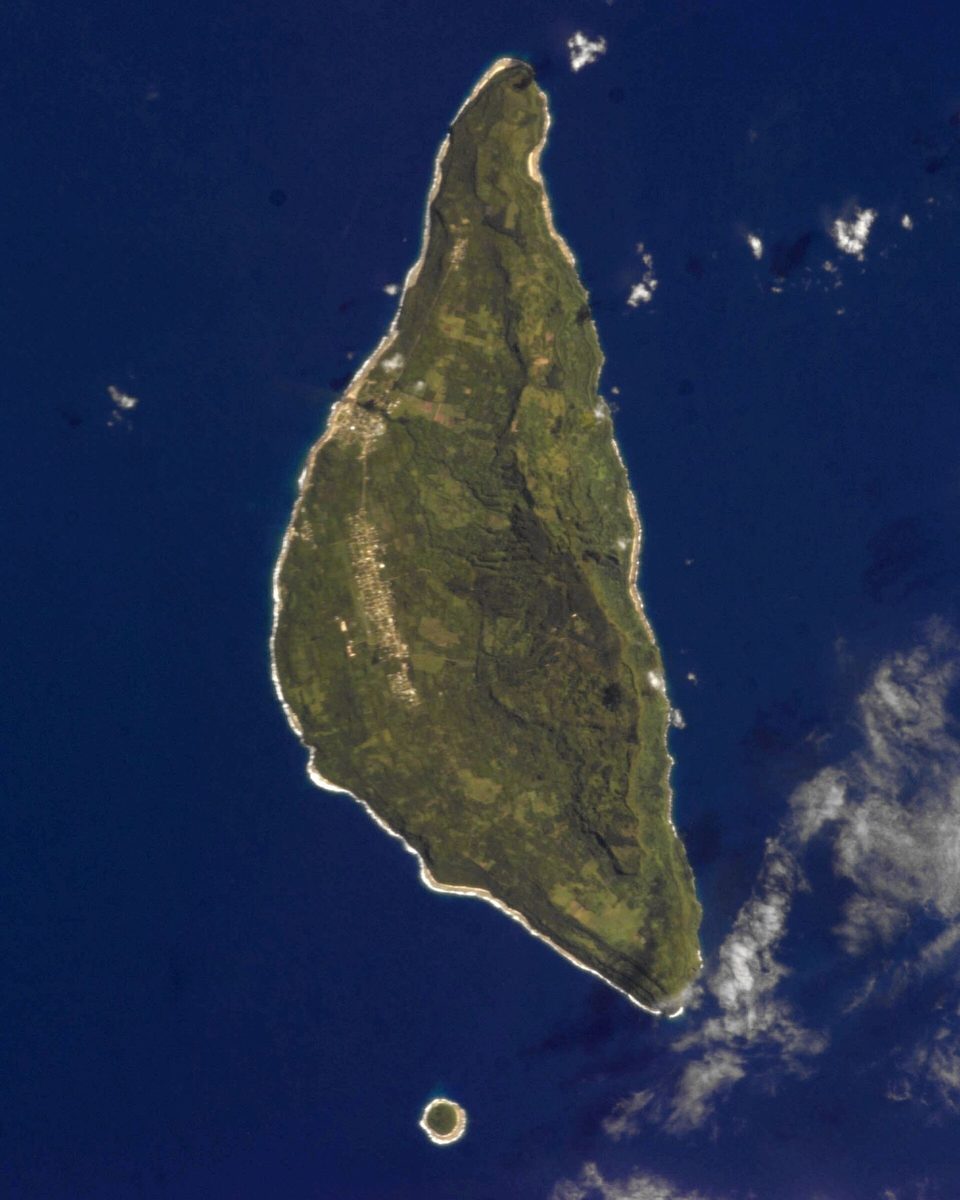 A long, tapering green island ringed by white surf in a deep-blue ocean, a small islet off its southern tip, seen from orbit