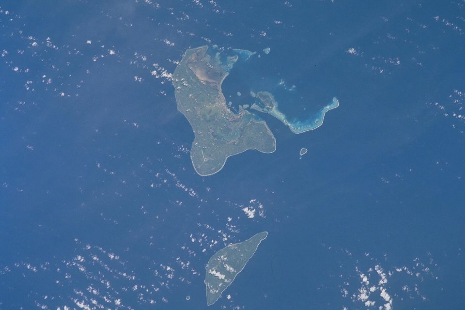 Two green islands in a deep-blue sea seen from orbit: a larger one with a pale lagoon and reefs above, a narrow one below