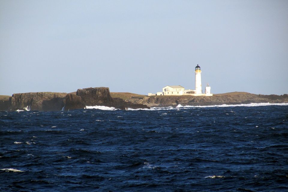 A white lighthouse tower and keepers' buildings stand on a low dark headland behind breaking surf and deep-blue choppy water